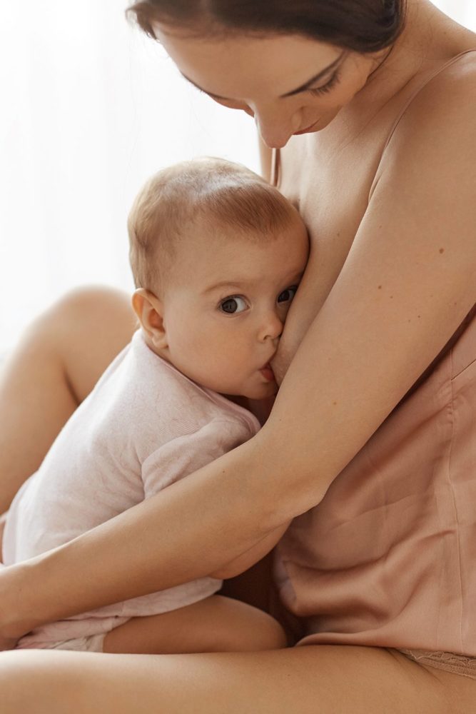 Young tender mother in sleepwear breastfeeding hugging her newborn baby sitting in bed at morning.